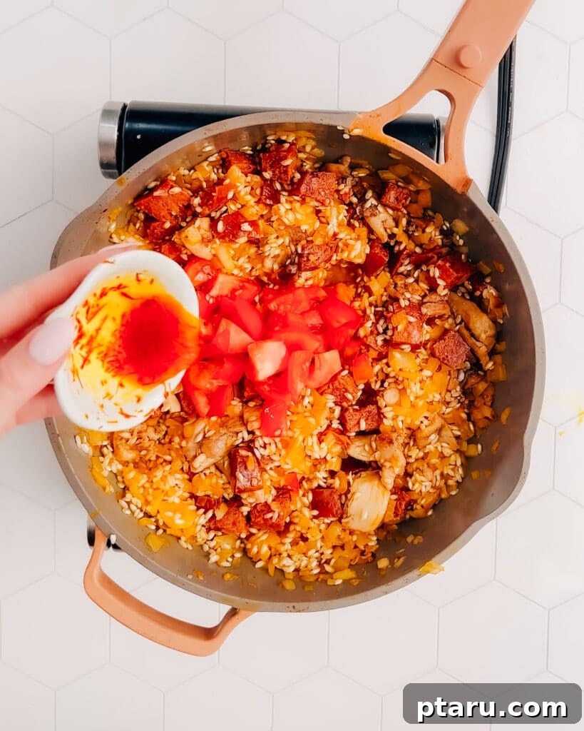 Spanish Surf and Turf Paella 5 Diced Roma tomatoes and saffron threads soaked in water being added to the paella base in a frying pan, enhancing color and flavor.