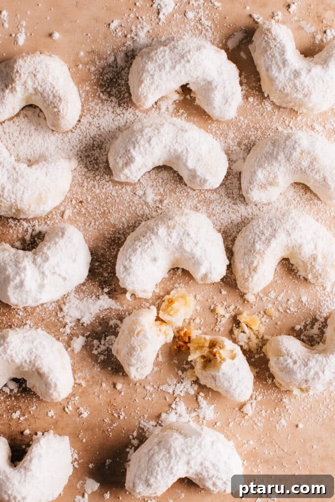 Kourabiedes Traditional Greek Christmas Butter Cookies 2 A batch of Kourabiedes or Greek Christmas Cookies on a surface with one cookie broken, revealing its delicate interior and almond pieces.
