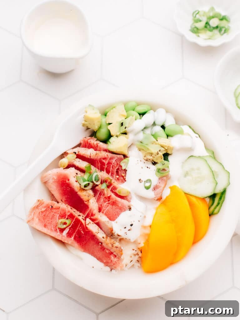 Effortless Air Fryer Tuna Steaks 2 A delicious air-fried tuna steak is served over a bed of rice, topped with dressing, cucumber, edamame beans, and sliced mango. A white spoon rests on the bowl, and small bowls of spring onions are placed nearby.