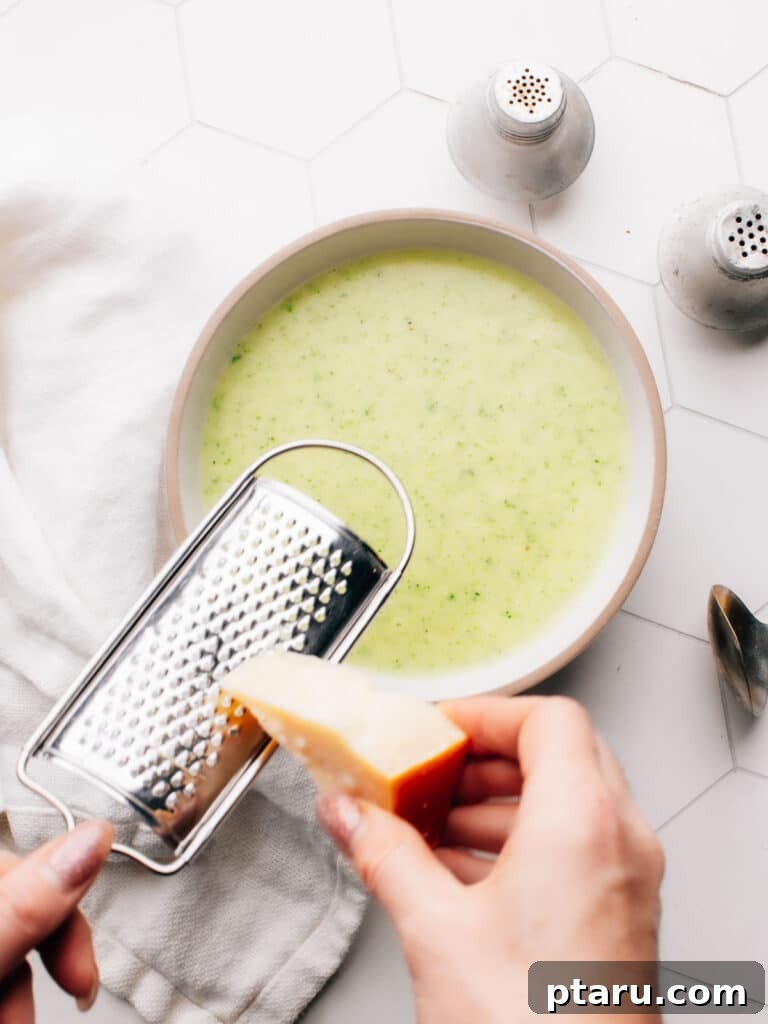 Silky Zucchini Potato Velouté 5 A person gracefully grating fresh Parmesan cheese over a steaming bowl of homemade potato zucchini soup, highlighting its comforting and gourmet appeal. The soup is ready to be enjoyed.