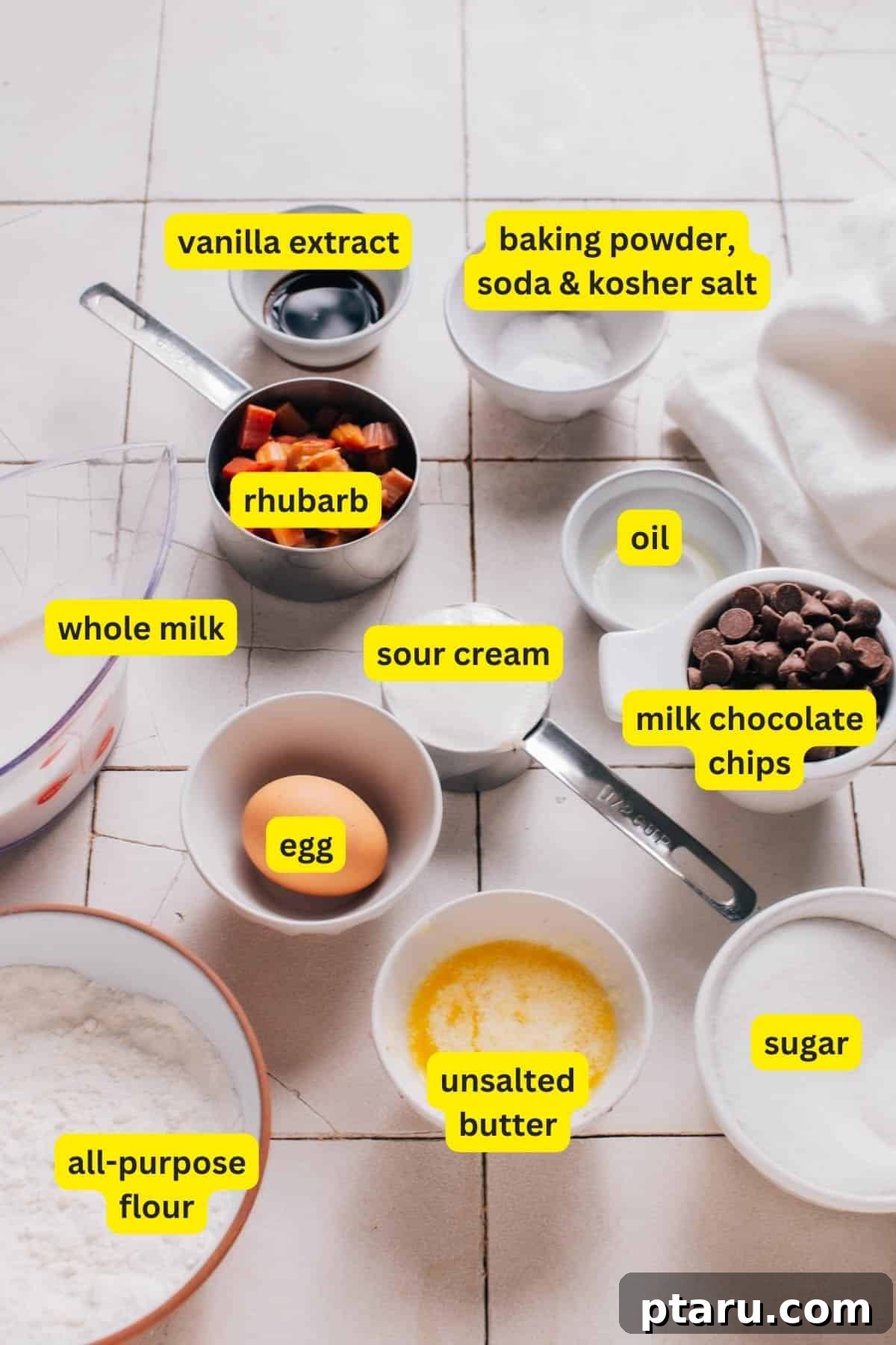 Tangy Rhubarb Delights 3 Ingredients for rhubarb cookies laid out on a kitchen countertop, including vanilla extract, baking powder, baking soda, kosher salt, rhubarb, whole milk, sour cream, oil, milk chocolate chips, all purpose flour, unsalted butter, sugar and egg.