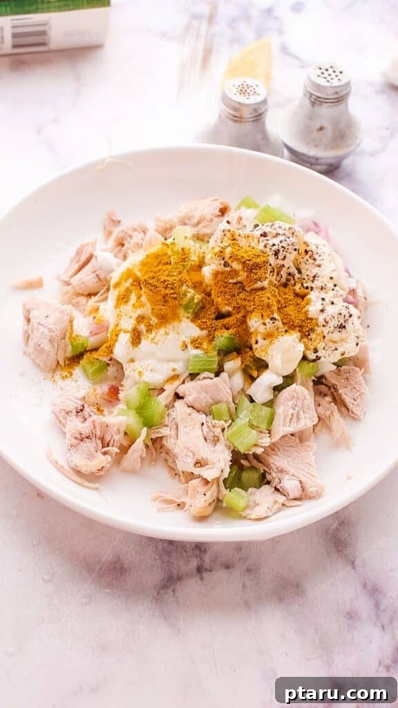 Flavorful Curried Chicken Salad with Sweet Grapes 7 A hand adding seasoning, including curry powder and salt, to a bowl of chicken salad for enhanced flavor.