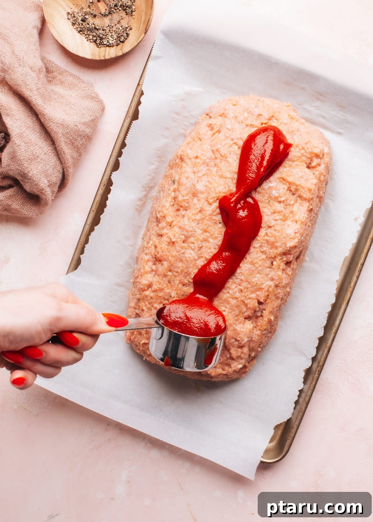 The Ultimate Juicy Turkey Meatloaf 6 A spoon spreading a rich, tangy sauce or glaze over a partially baked turkey meatloaf on a baking sheet.