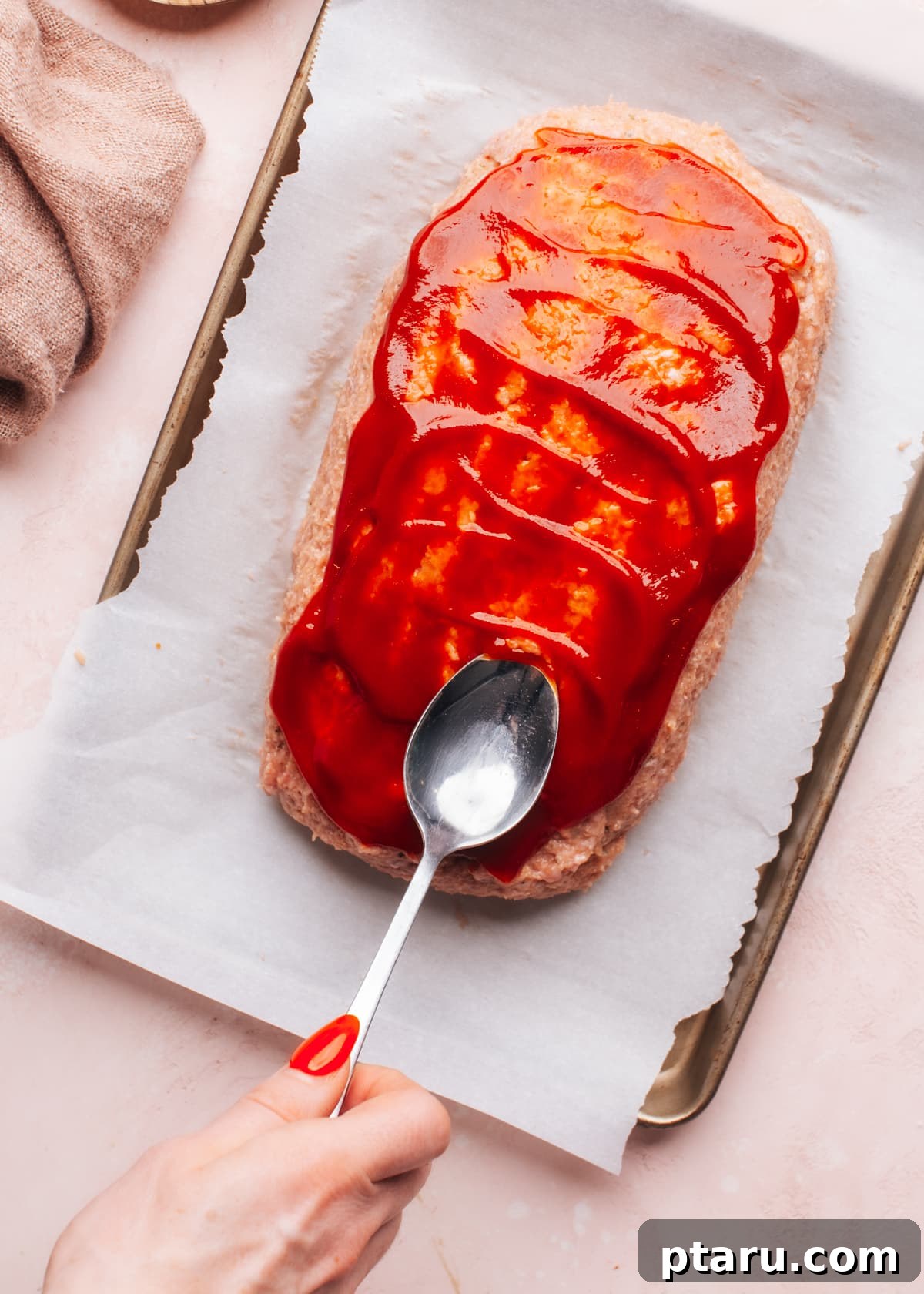 The Ultimate Juicy Turkey Meatloaf 7 A hand gently spreading a vibrant sauce over a baked meatloaf with a spoon, enhancing its appearance and flavor.