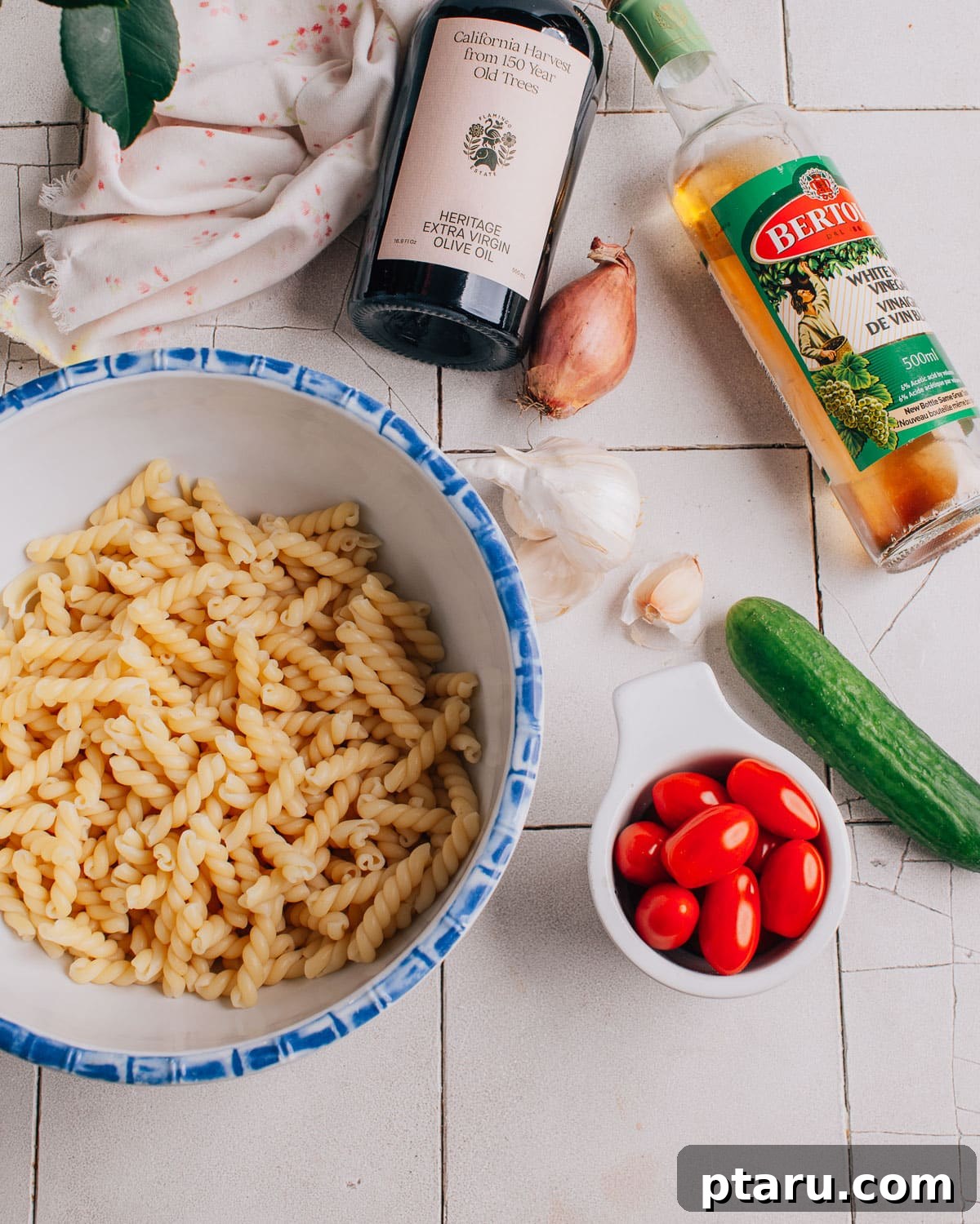 Tuscan Pasta Toss 3 A flat lay of fresh ingredients for Pasta Salad with Italian Dressing, including uncooked pasta, cherry tomatoes, cucumber, and a bowl of dressing.
