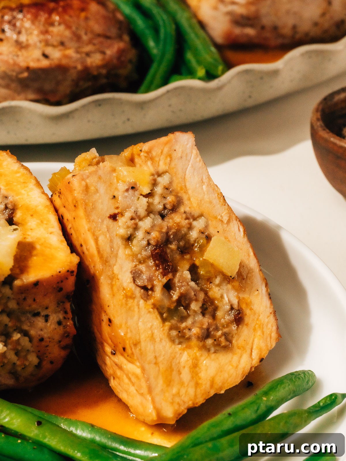 Juicy Stuffed Pork Chops with Luscious Pan Sauce 2 A beautifully cooked stuffed pork chop on a plate, cut open to reveal the flavorful apple and sausage filling, with more golden-brown stuffed pork chops and a vibrant green side dish blurred in the background.