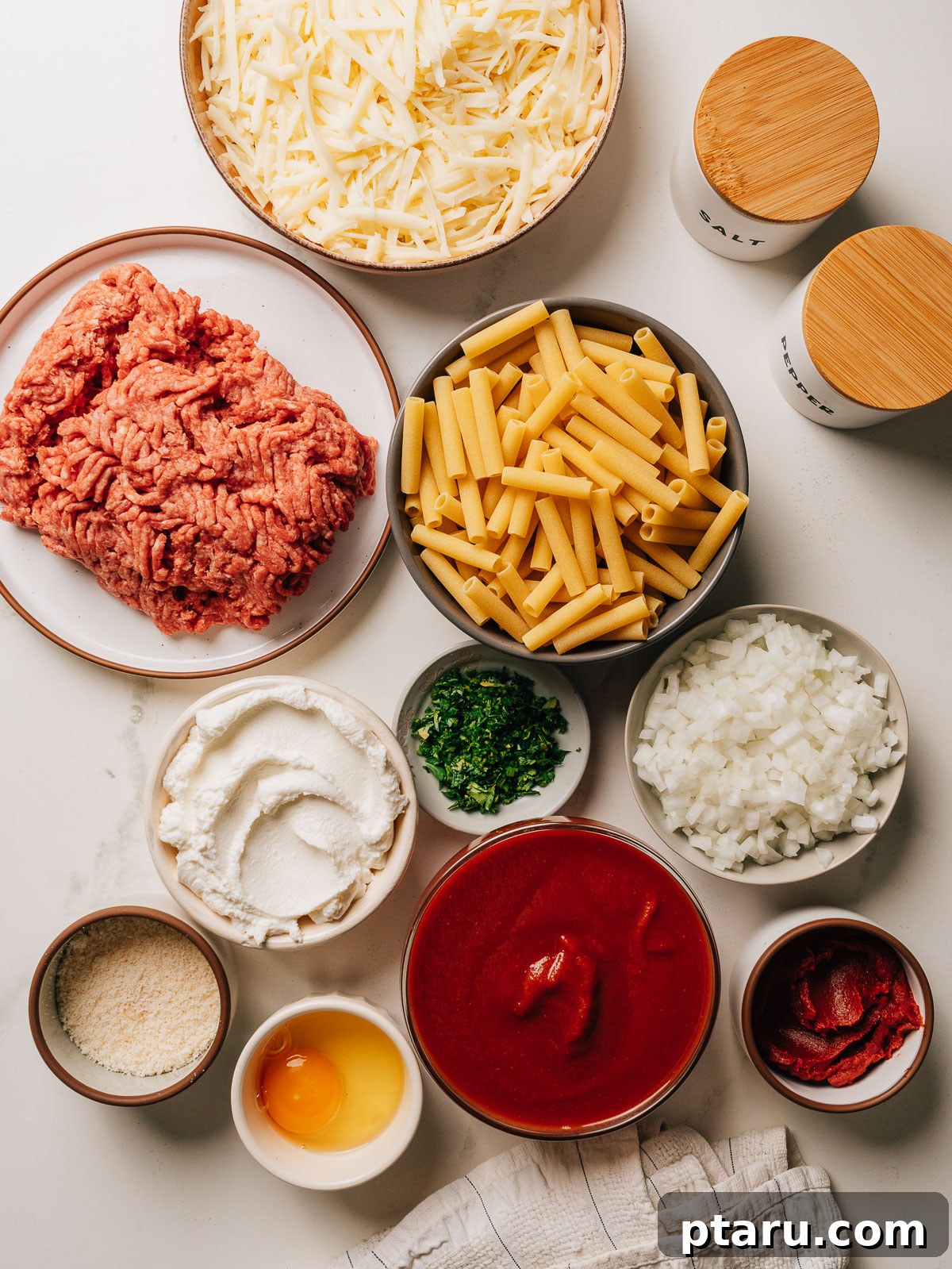 Ultimate Ricotta Ziti Bake 3 Ingredients for baked ziti including ziti pasta, ground beef, ricotta, mozzarella, tomatoes, and fresh herbs laid out on a table.