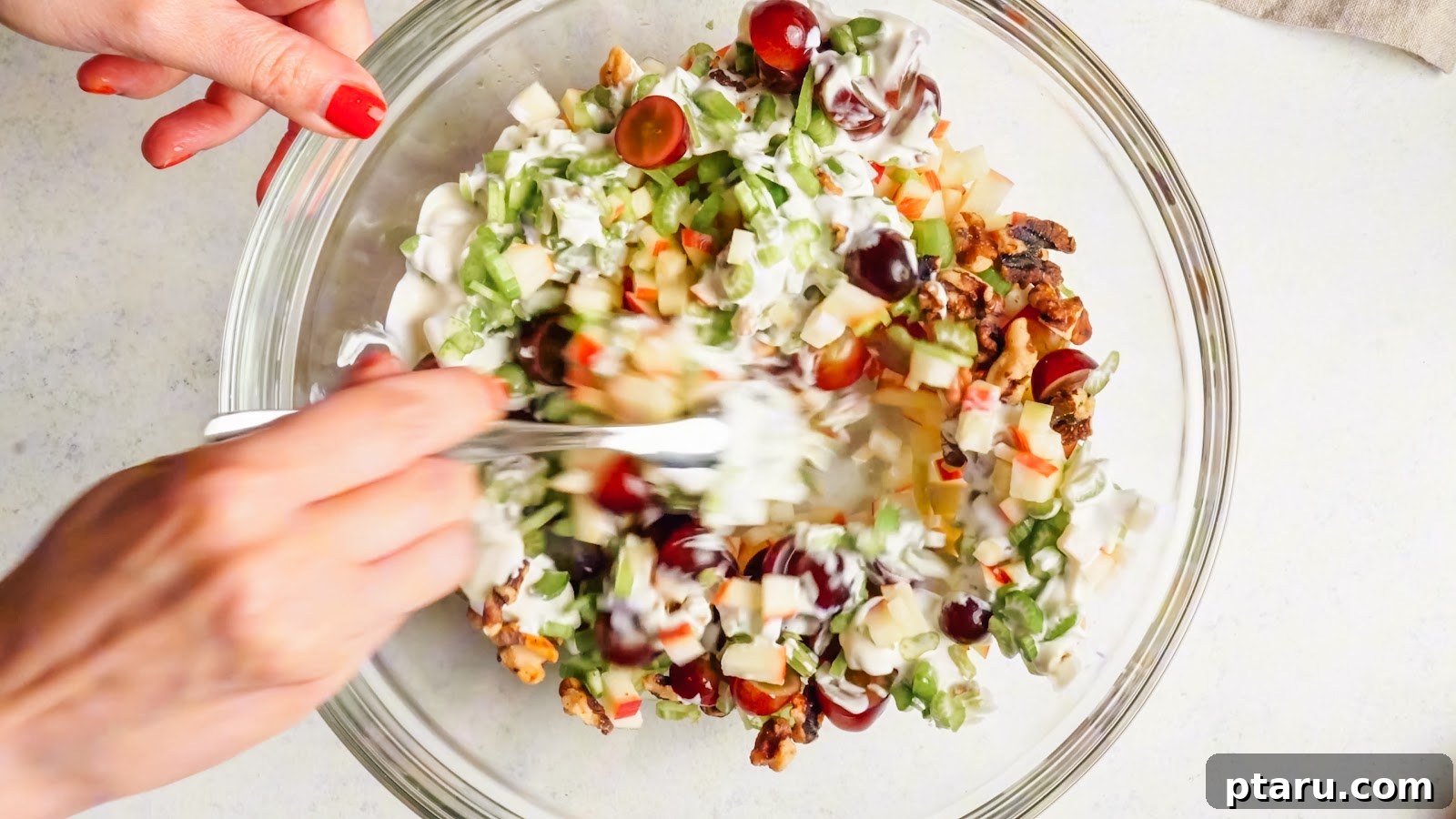 Classic Waldorf Salad with Apples, Grapes, and Walnuts 8 Folding the dressing through the Waldorf salad with a fork in a glass bowl.