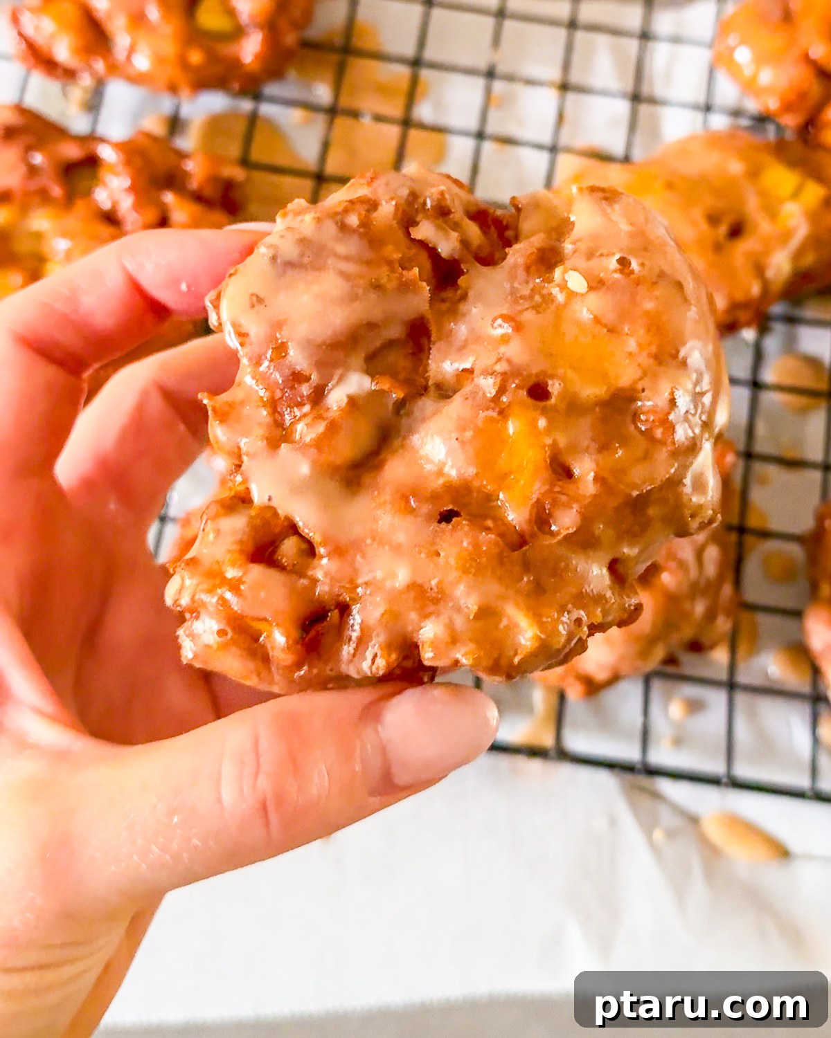 Crispy Homemade Apple Fritters with Cinnamon Glaze 2 A hand holding a glazed apple fritter over a wire rack.